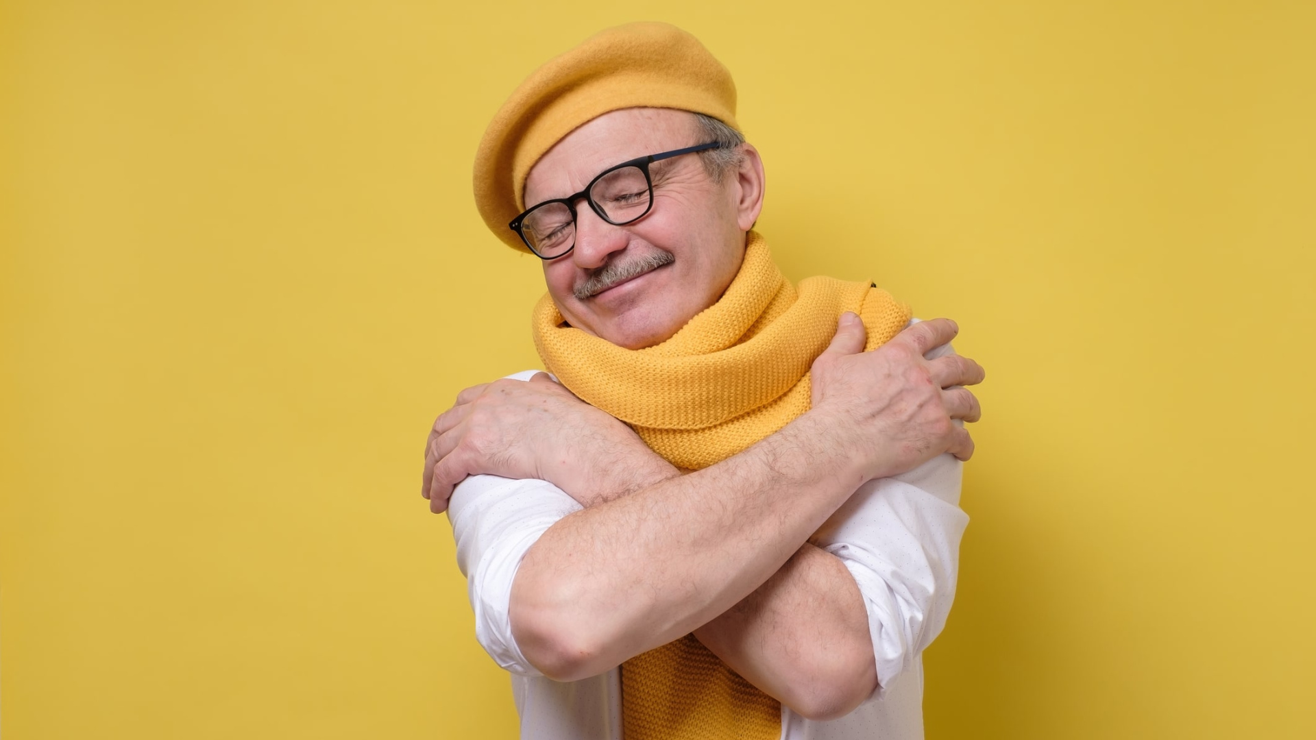 older man hugging himself happily wearing knitted hat and scarf